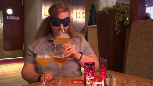 Eine Frau mit einer blickdichten Augenmaske testet drei unterschiedliche Biere.