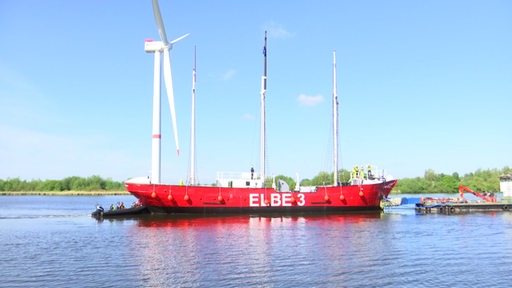 Das Museumsschiff "Elbe 3" schippert auf einem Gewässer. Im Hintergrund sieht man den blauen Himmel und ein Windrad.