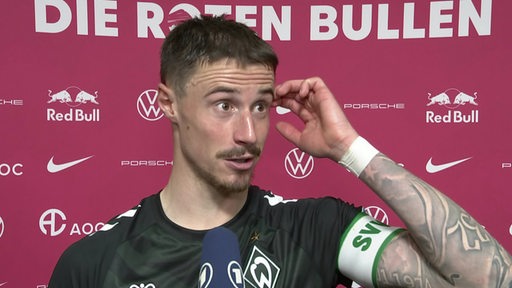 Werder-Kapitän Marco Friedl steht vor einer pinkfarbenen Werbewand beim Interview nach dem Spiel in Leipzig.
