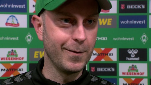 Werder-Trainer Ole Werner steht vor einer Werbewand und lächelt beim Interview nach dem Spiel.