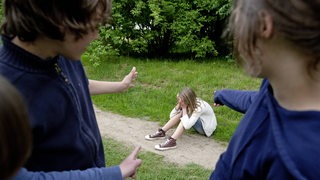 Ein Mädchen sitzt auf dem Boden und wird von Mitschülern gemobbt (gestellte Szene) 