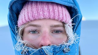 Eine junge Frau mit Kapuze auf dem Kopf blickt mit gefrorenen Haaren und Wimpern in die Kamera
