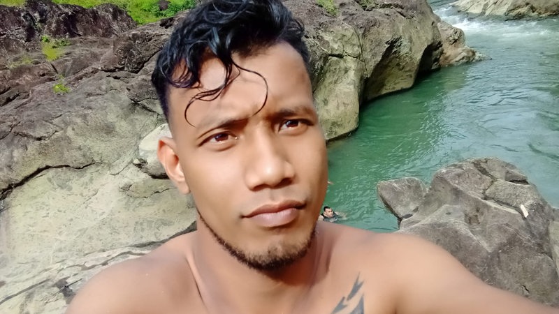 Ein Mann mit nacktem Oberkörper und Tattoo macht ein Selfie vor Felsen und Wasser.