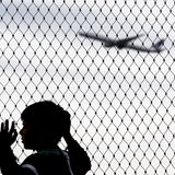 Ein Kind steht an einem hohen Zaun, am Himmel ein Flugzeug