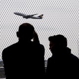 Familie von hinten sieht einem wegfliegenden Flugzeug durch einen Zaun nach.