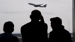 Familie von hinten sieht einem wegfliegenden Flugzeug durch einen Zaun nach.