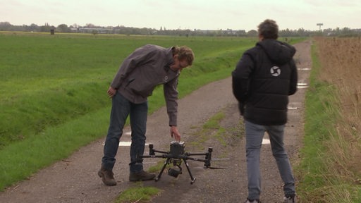 Auf einem Feldweg zwischen Feldern sind zwei Männer mit einer Drohne zu sehen.