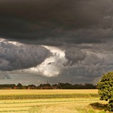 Dunkle Wolken hängen über einem Acker. Im Hintergrund ist eine Ortschaft zu sehen.