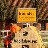 Ein Ortsschild mit der Aufschrift "Blender" Ortsteil Adolfshausen Landkreis Verden ist zu sehen. Davor steht ein Stein mit der Aufschrift "Adolfshausen" und darüber ist eine Krone eingeprägt.