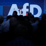 Delegierte sitzen vor dem Parteilogo bei dem AfD-Bundesparteitag.