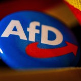 Ein Ansteckpin mit dem Parteilogo der AfD liegt mit einer Deutschland-Banderole auf einem Tisch.