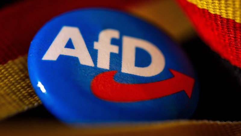 Ein Ansteckpin mit dem Parteilogo der AfD liegt mit einer Deutschland-Banderole auf einem Tisch.