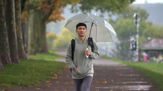 Ein Mann mit einem hellen Regenschirm geht eine Allee mit Bäumen entlang und blickt nach vorn in die Kamera.