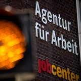 Eine Ampel leuchtet vor dem Schild der Agentur für Arbeit und des Jobcenter.