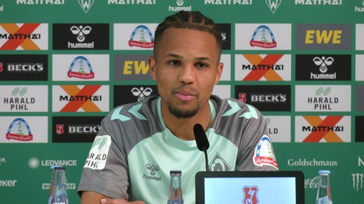 Werderspieler Felix Agu sitzt im Trikot vor einem Mikrofon während einer Pressekonferenz im Weserstadion.