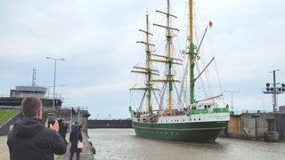 Mehrere Menschen stehen am Wasser und beobachten ein einlaufendes Segelschiff.