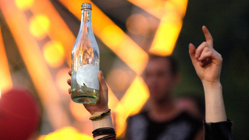 Zwei Hände zeigen in die Luft, eine davon hält eine leere Flasche Alkohol.