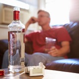 Auf einem Wohnzimmertisch steht eine Flasche Schnaps, im Hintergrund ein Mann mit Glas auf dem Sofa.