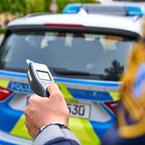 Ein Polizist steht hinter einem Streifenwagen und hält ein Alkoholmessgerät in der Hand.