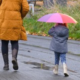 Eine Frau, die einen Kinderrucksack trägt, und ein Kind, das einen aufgespannten Regenschirm über dem Kopf hält, gehen eine mit Pfützen versehene Straße entlang.