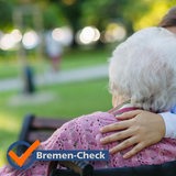 Eine Frau in blauer Pflegekleidung legt den Arm um eine Seniorin im Rollstuhl.