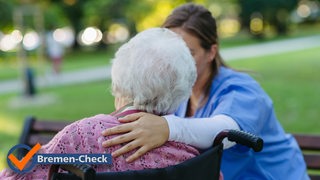 Eine Frau in blauer Pflegekleidung legt den Arm um eine Seniorin im Rollstuhl.