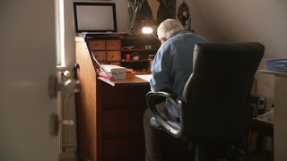 Ein älterer Mann, der an einem Schreibtisch sitzt und der Kamera den Rücken zugewandt hat.