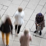 An einer  alten Frau mit einer Krücke laufen (bewegungsunscharf) Menschen vorbei.