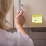 Eine alzheimerkranke Frau öffnet einen Küchenschrank, auf dem ein Hinweis "Herd aus?" steht (Symbolbild)