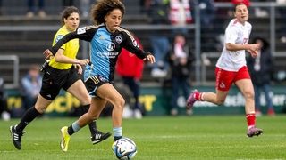 Die Fußballspielerin Amira Arfaoui sprintet mit dem Ball am Fuß über das Spielfeld. Im Hintergrund ist die Schiedsrichterin und eine Gegenspielerin zu sehen.