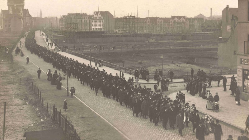 Ein schwarz-weiß Foto von einem langen Trauerzug mit vielen schwarz gekleideten Menschen.