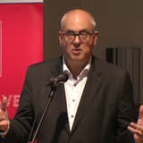 Ein Mann steht gestekulierend an einem Mikrofon.