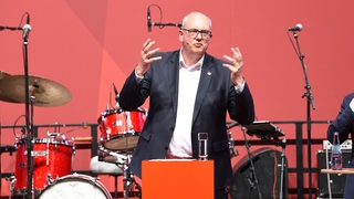Ein Mann im Anzug und mit Brille steht auf einer Bühne. Hinter ihm ein Schlagzeug, vor ihm ein Pult mit der Aufschrift SPD.