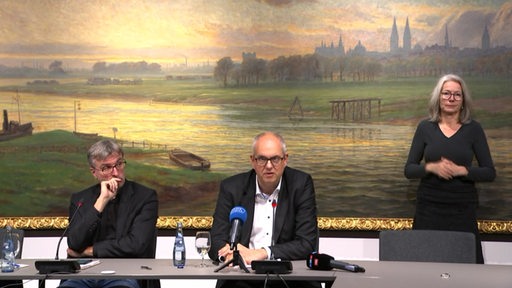 Bürgermeister Andreas Bovenschulte spricht auf einer Pressekonferenz, neben ihm Pressesprecher Christian Dohle und eine Gebärdendolmetscherin.
