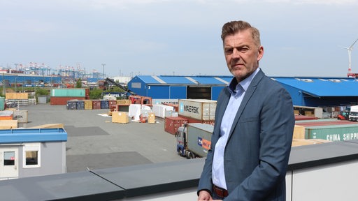 Ein Mann steht vor einem Hafen-Panorama mit Hallen, Containern und Kränen.