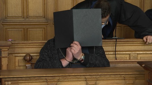 Ein Angeklagter sitzt auf der Gerichtsbank und verdeckt sein Gesicht mit einem schwarzen Schnellhefter.