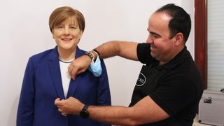 Eine Figur, die aussieht wie Angela Merkel, steht vor einer weißen Wand.