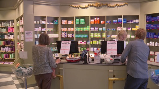In einer Apotheke stehen zwei Kunden und im Hintergrund ist ein großes Regal mit unterschiedlichen Arzneimitteln zu sehen.