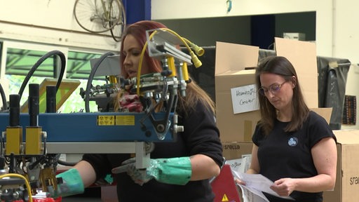 Zwei Frauen auf der Arbeit. Eine der Frauen an einer Maschine, die andere mit Papier in der Hand.