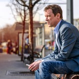 Ein Mann sitzt mit Handy in der Hand auf einer Bank und schaut nachdenklich.