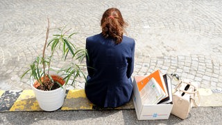 Eine Arbeitnehmerin, der gekündigt wurde, sitzt mit ihren Kartons und Pflanzen auf einem Bordstein