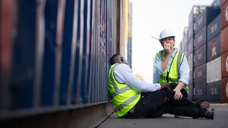 Eine Frau sitzt neben einem Mann, dem es offenbar nicht gut geht. Sie ruft etwas in ein Walkie-Talkie hinein.
