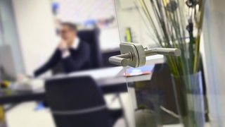  Blick in durch eine offene Glastür in einem Büro. Ein Mann sitzt an seinem Schreibtisch (Symbolbild)