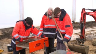 Zwei Mitarbeiter und eine Mitarbeiterin der Landesarchäologie Bremen stehen in einem Zelt und sieben mit einem Gerät Erde.