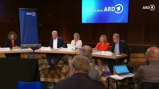 Vier Personen an einem Tisch sitzend. Im Hintergrund das ARD-Logo.