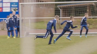Zwei Jungen in Sportklamotten des Leher TS rennen über einen Fußballplatz.