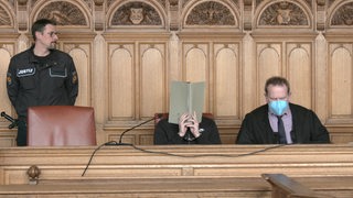 Der Angeklagte sitzt vor dem Gericht neben ihm sein Anwalt und ein Polizist. 