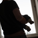 Eine Person von hinten, die ein Kind auf dem Arm hält. Es ist dunkel.