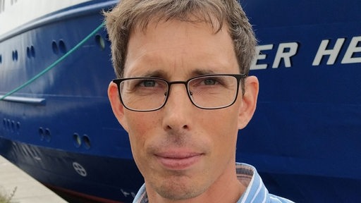 Ein Mann mit Brille steht vor einem Schiff und guckt in die Kamera.