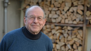 Älterer Herr mit blauem Pulli und Brille blickt vor Holzscheiteln im Hintergrund in die Kamera